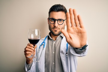 Young doctor man wearing stethoscope drinking a glass of fresh wine over isolated background with open hand doing stop sign with serious and confident expression, defense gesture