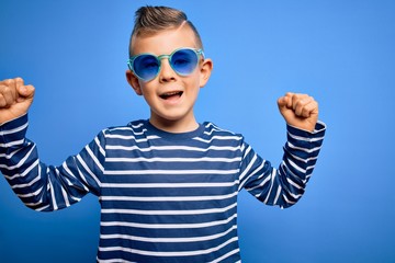 Young little caucasian kid with blue eyes standing wearing sunglasses over blue background celebrating surprised and amazed for success with arms raised and open eyes. Winner concept.