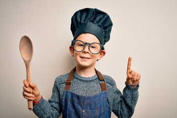Young little caucasian cook kid wearing chef uniform and hat cooking using wooden spoon surprised...