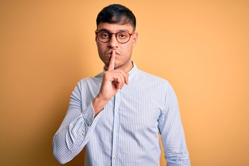 Young handsome hispanic business man wearing nerd glasses over yellow background asking to be quiet with finger on lips. Silence and secret concept.