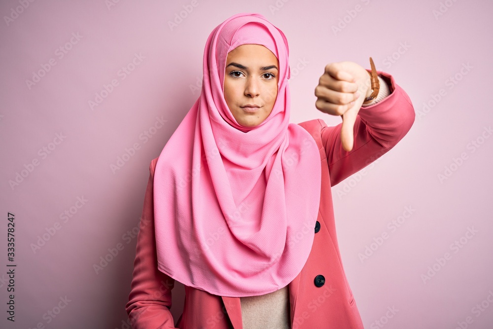 Wall mural Young beautiful girl wearing muslim hijab standing over isolated pink background looking unhappy and angry showing rejection and negative with thumbs down gesture. Bad expression.