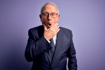 Grey haired senior business man wearing glasses and elegant suit and tie over purple background Looking fascinated with disbelief, surprise and amazed expression with hands on chin
