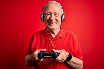 Senior grey haired gamer man playing video games using gamepad joystick over red background with a happy face standing and smiling with a confident smile showing teeth