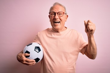 Grey haired senior football player man holding soccer ball over pink isolated background screaming proud and celebrating victory and success very excited, cheering emotion