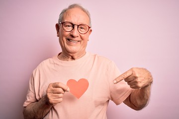 Senior grey haired man holding heart shape paper over pink background very happy pointing with hand and finger