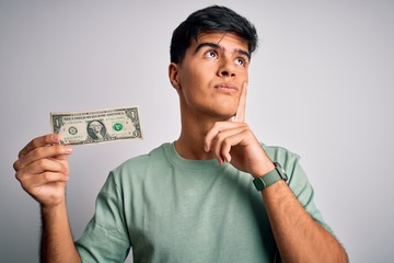 Young handsome man holding one dollar banknotes over isolated white background serious face...