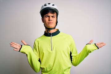 Young handsome cyclist man wearing security bike helmet over isolated white background clueless and confused expression with arms and hands raised. Doubt concept.