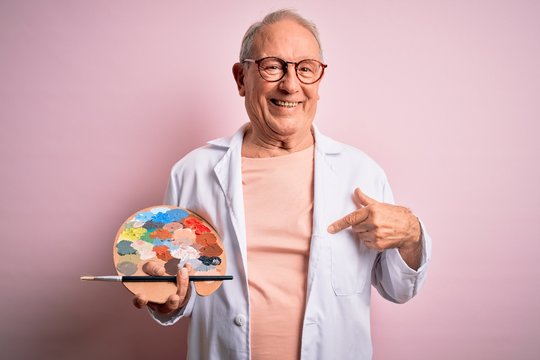 Senior grey haired artist man painting using painter palette over pink background with surprise face pointing finger to himself