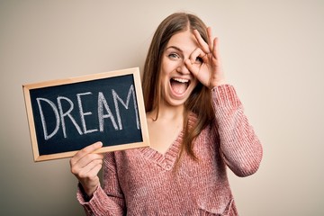 Young beautiful redhead woman holding blackboard with dream word message with happy face smiling doing ok sign with hand on eye looking through fingers