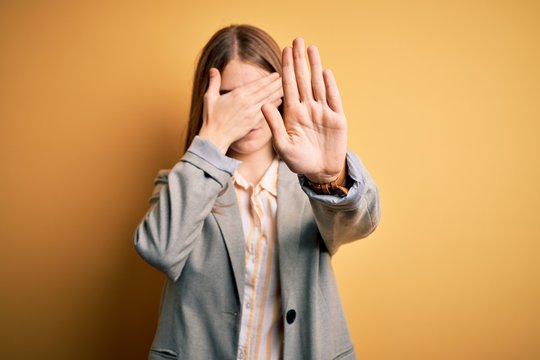 Young beautiful redhead woman wearing jacket and glasses over isolated yellow background covering eyes with hands and doing stop gesture with sad and fear expression. Embarrassed and negative concept.