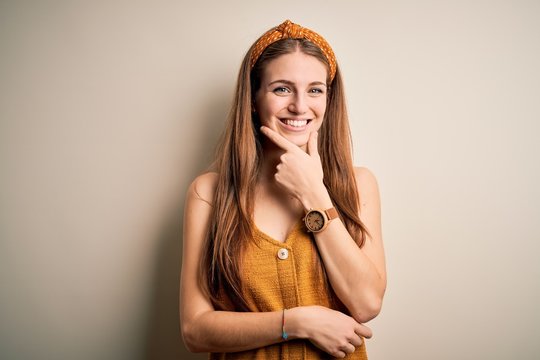 Young beautiful redhead woman wearing casual t-shirt and diadem over yellow background looking confident at the camera smiling with crossed arms and hand raised on chin. Thinking positive.