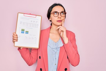 Young beautiful business woman with blue eyes wearing glasses holding clipboard serious face...
