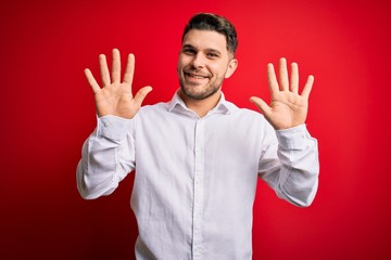 Young business man with blue eyes wearing elegant shirt standing over red isolated background showing and pointing up with fingers number ten while smiling confident and happy.