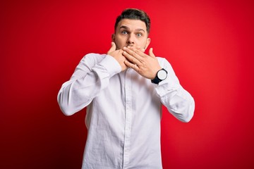 Young business man with blue eyes wearing elegant shirt standing over red isolated background shocked covering mouth with hands for mistake. Secret concept.