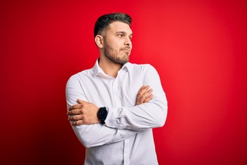 Young business man with blue eyes wearing elegant shirt standing over red isolated background looking to the side with arms crossed convinced and confident