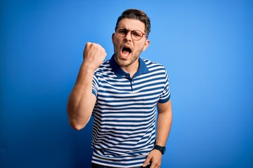 Young man with blue eyes wearing glasses and casual striped t-shirt over blue background angry and mad raising fist frustrated and furious while shouting with anger. Rage and aggressive concept.