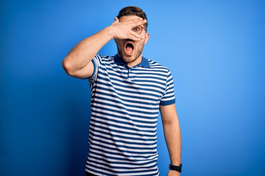 Young man with blue eyes wearing glasses and casual striped t-shirt over blue background peeking in shock covering face and eyes with hand, looking through fingers with embarrassed expression.