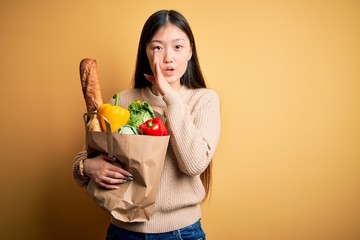 Young asian woman holding paper bag of fresh healthy groceries over yellow isolated background hand on mouth telling secret rumor, whispering malicious talk conversation