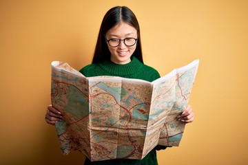 Young asian turist woman looking at city tourist map on a trip over yellow background with a happy face standing and smiling with a confident smile showing teeth