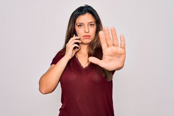 Young hispanic woman having a conversation talking on smartphone over isolated background with open hand doing stop sign with serious and confident expression, defense gesture