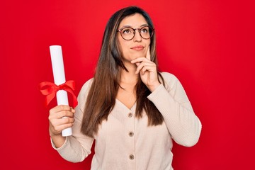 Young hispanic smart woman wearing glasses holding university degree over red background serious face thinking about question, very confused idea