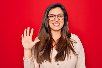 Young hispanic smart woman wearing glasses standing over red isolated background showing and pointing up with fingers number five while smiling confident and happy.