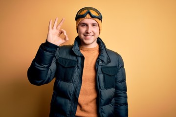 Young handsome caucasian man wearing hat, coat and ski glasses for winter and snow weather smiling positive doing ok sign with hand and fingers. Successful expression.