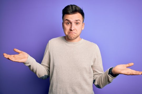 Young handsome caucasian man wearing casual sweater over purple isolated background clueless and confused expression with arms and hands raised. Doubt concept.