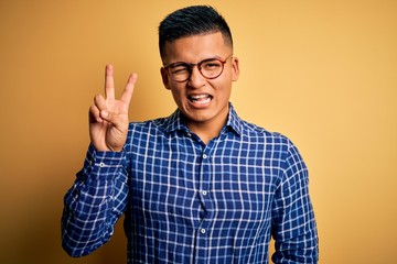 Naklejka premium Young handsome latin man wearing casual shirt and glasses over yellow background smiling with happy face winking at the camera doing victory sign with fingers. Number two.