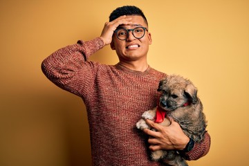 Young handsome latin man holding cute puppy pet over isolated yellow background stressed with hand...