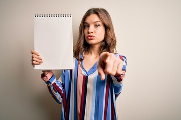 Young blonde girl showing white space paper on notebook over isolated background pointing with finger to the camera and to you, hand sign, positive and confident gesture from the front