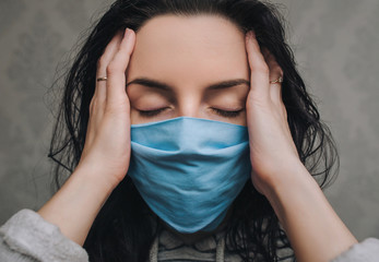 A young girl in a blue protective medical mask respirator is holding head in hands on a gray wall...