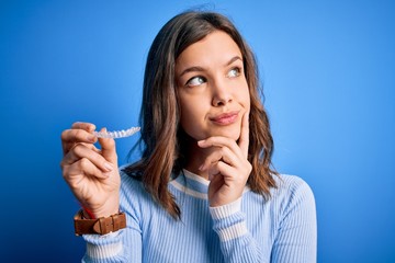 Young blonde girl holding dental orthodontic clear aligner over blue isolated background serious face thinking about question, very confused idea