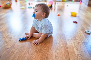 Adorable toddler sitting on the floor using pacifier playing around lots of toys at kindergarten
