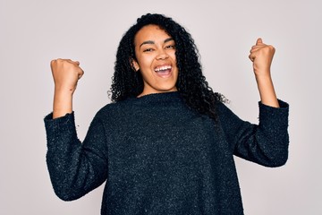 Young african american curly woman wearing casual sweater standing over white background celebrating surprised and amazed for success with arms raised and open eyes. Winner concept.