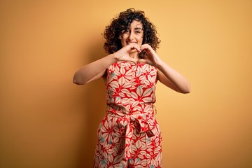 Young beautiful curly arab woman on vacation wearing summer floral dress and sunglasses smiling in love doing heart symbol shape with hands. Romantic concept.
