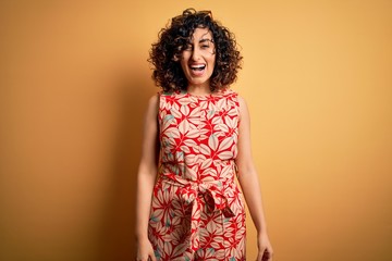 Young beautiful curly arab woman on vacation wearing summer floral dress and sunglasses winking...