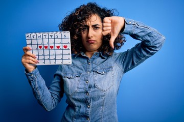 Young beautiful curly arab woman holding period calendar controlling menstrual cycle with angry...