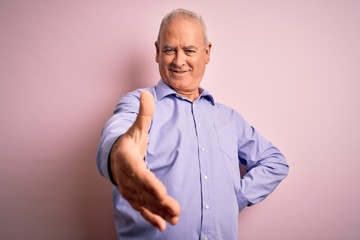 Middle age handsome hoary man wearing casual shirt standing over pink background smiling friendly offering handshake as greeting and welcoming. Successful business.