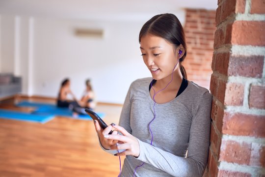 Young beautiful chinese sportswoman smiling happy. Standing listening to music using earphones and smartphone after class of yoga at gym