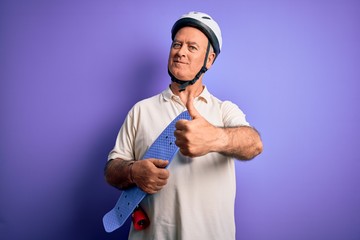 Naklejka premium Middle age hoary man wearing security helmet holding skateboard over purple background happy with big smile doing ok sign, thumb up with fingers, excellent sign