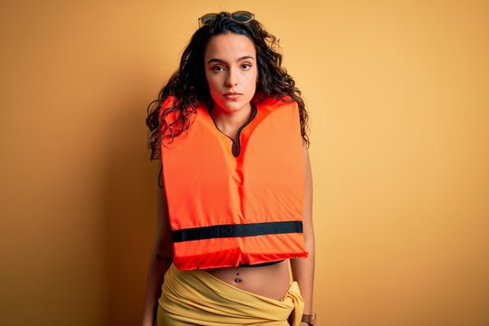 Young beautiful woman with curly hair wearing orange lifejacket over yellow background with serious expression on face. Simple and natural looking at the camera.