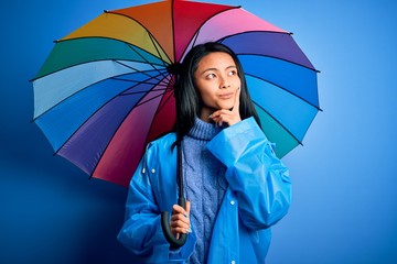 Young chinese woman wearing rain coat holding umbrella over isolated blue background serious face...