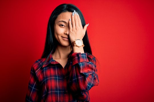 Young beautiful chinese woman wearing casual shirt over isolated red background covering one eye with hand, confident smile on face and surprise emotion.