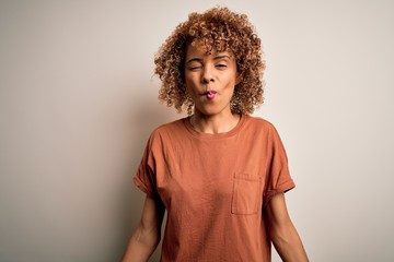 Beautiful african american woman with curly hair wearing casual t-shirt over white background making fish face with lips, crazy and comical gesture. Funny expression.