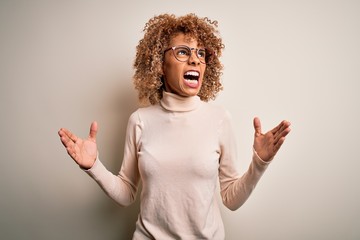 Young african american woman wearing turtleneck sweater and glasses over white background crazy and mad shouting and yelling with aggressive expression and arms raised. Frustration concept.