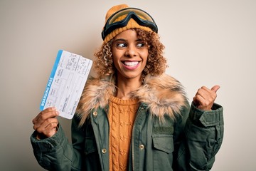 Young african american tourist woman wearing ski goggles holding plane boarding pass tickets pointing and showing with thumb up to the side with happy face smiling