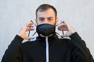 Brunette guy in a black tracksuit puts on a face mask. Black stylish face mask during a pandemic virus crown. Handmade cotton face mask for protection against viruses and bacteria.