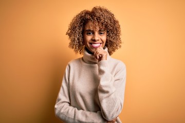 Obraz na płótnie Canvas Young beautiful african american woman wearing turtleneck sweater over yellow background looking confident at the camera smiling with crossed arms and hand raised on chin. Thinking positive.