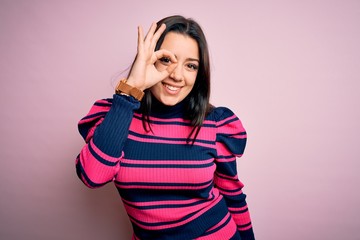 Obraz na płótnie Canvas Young brunette elegant woman wearing striped shirt over pink isolated background doing ok gesture with hand smiling, eye looking through fingers with happy face.